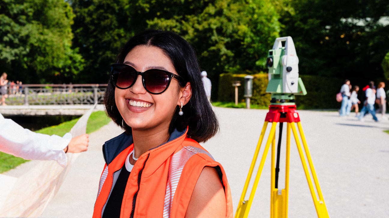 Das Foto zeigt eine Frau mit Sonnenbrille, die in einem Park steht. Im Hintergrund ist ein Tachymeter zu sehen.