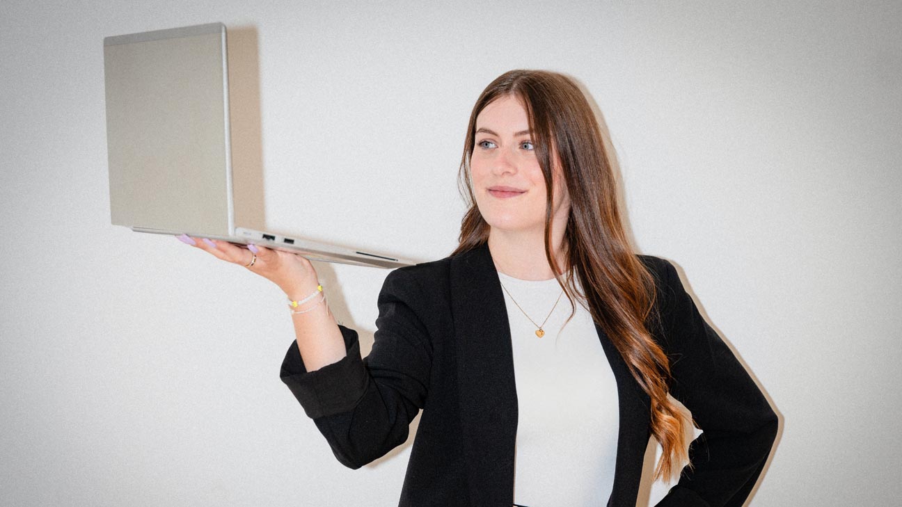 Auf dem Foto ist eine Frau mit einem Laptop in der Hand und zwei Männer leicht hinter ihr abgelichtet, die die Arme vor der Brust verschränken. Die drei Personen stehen in einem Verwaltungsgebäude, im Hintergrund ist eine Glasfront zu sehen.